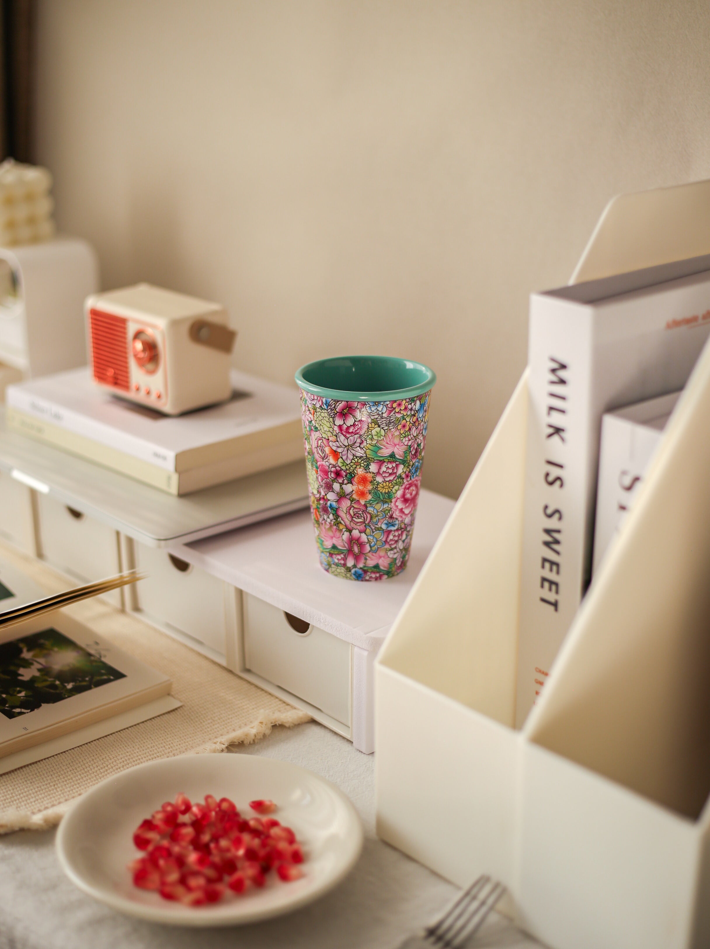 Colorful floral-patterned Falangcai cup on a shelf with books and a small dish of pomegranate seeds.