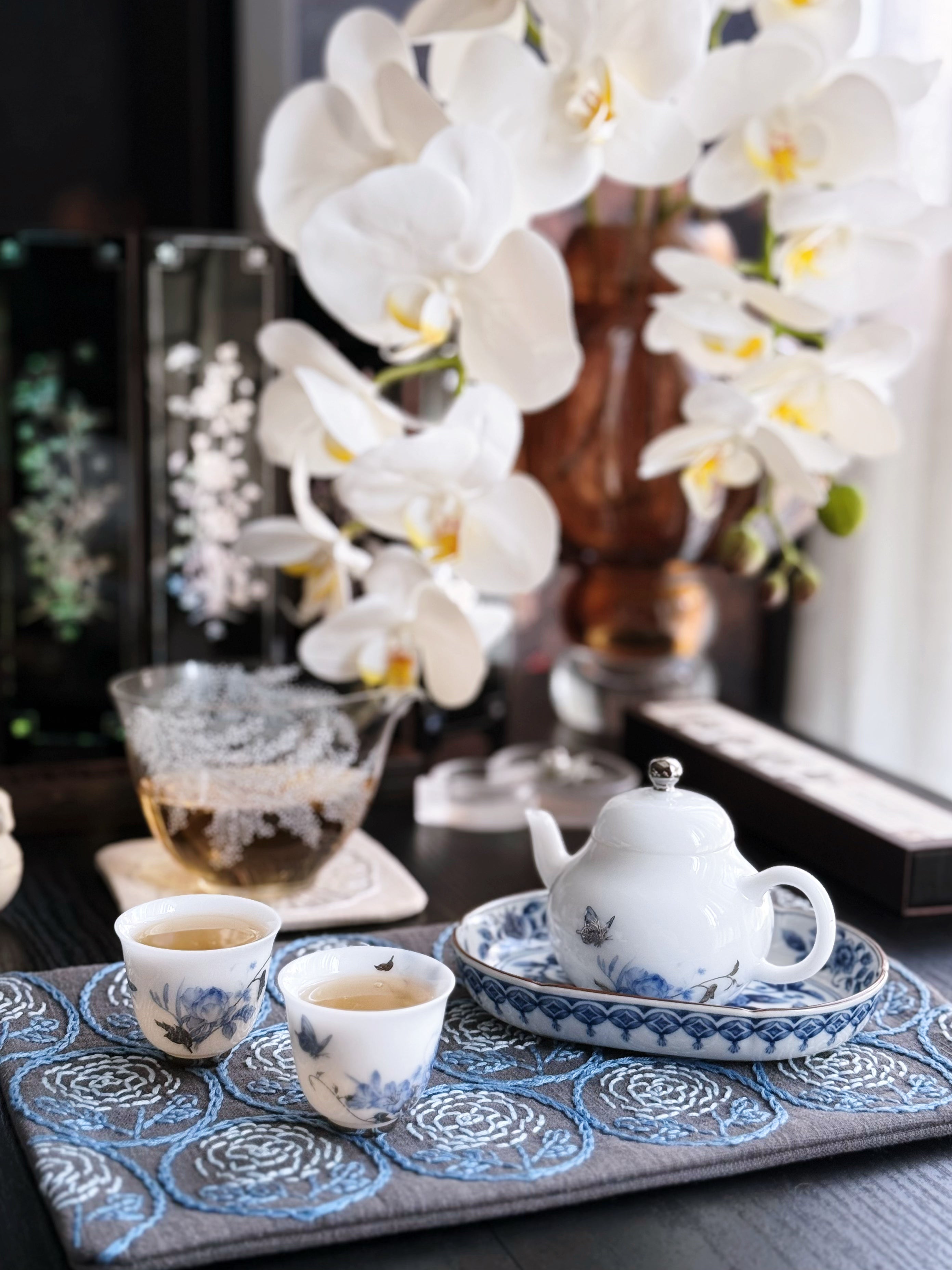 Tea set with teapot and cups on a decorative mat, accompanied by white orchids.