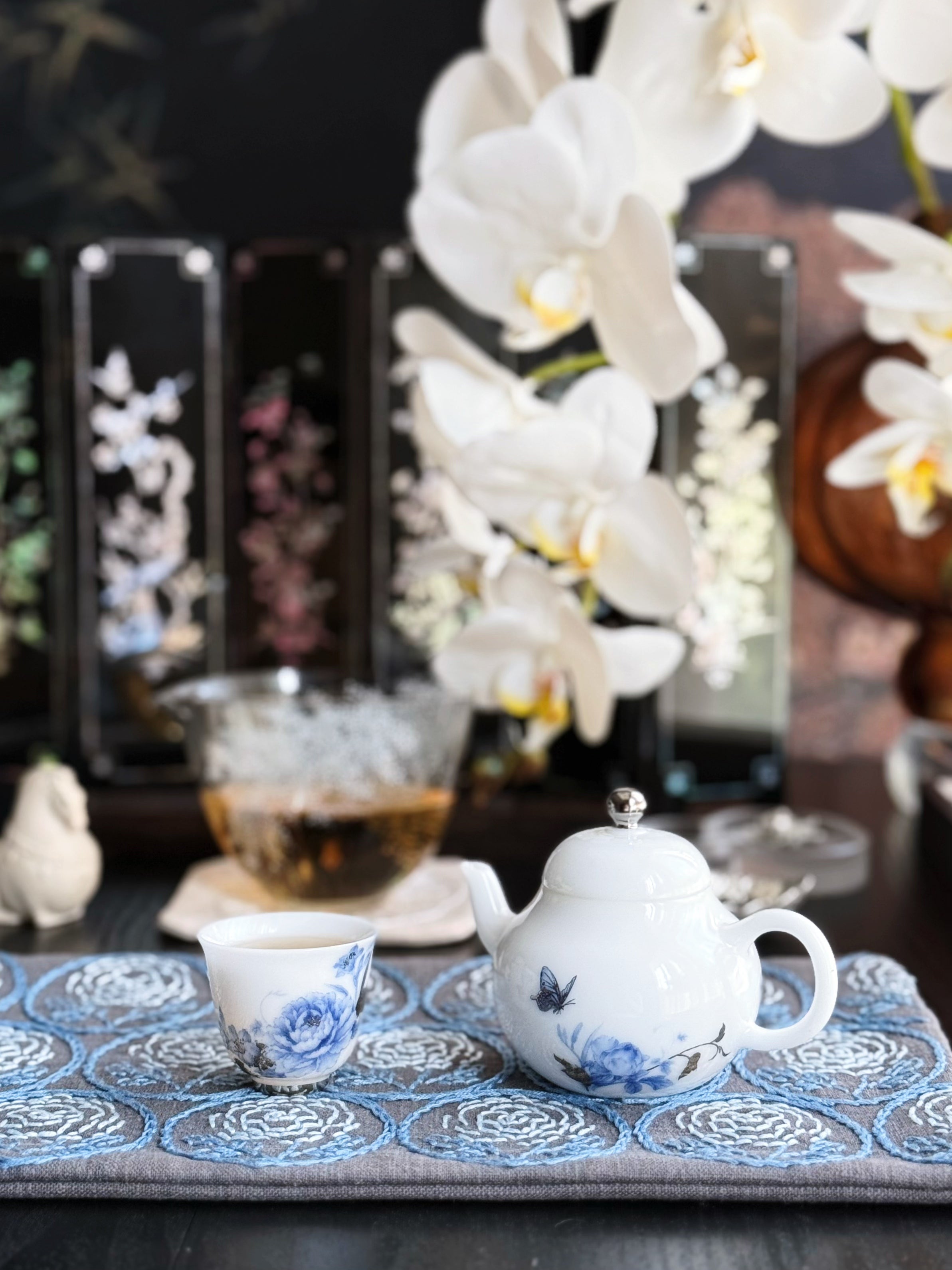 Tea set with a teapot and cup on a decorative mat, surrounded by white orchids.
