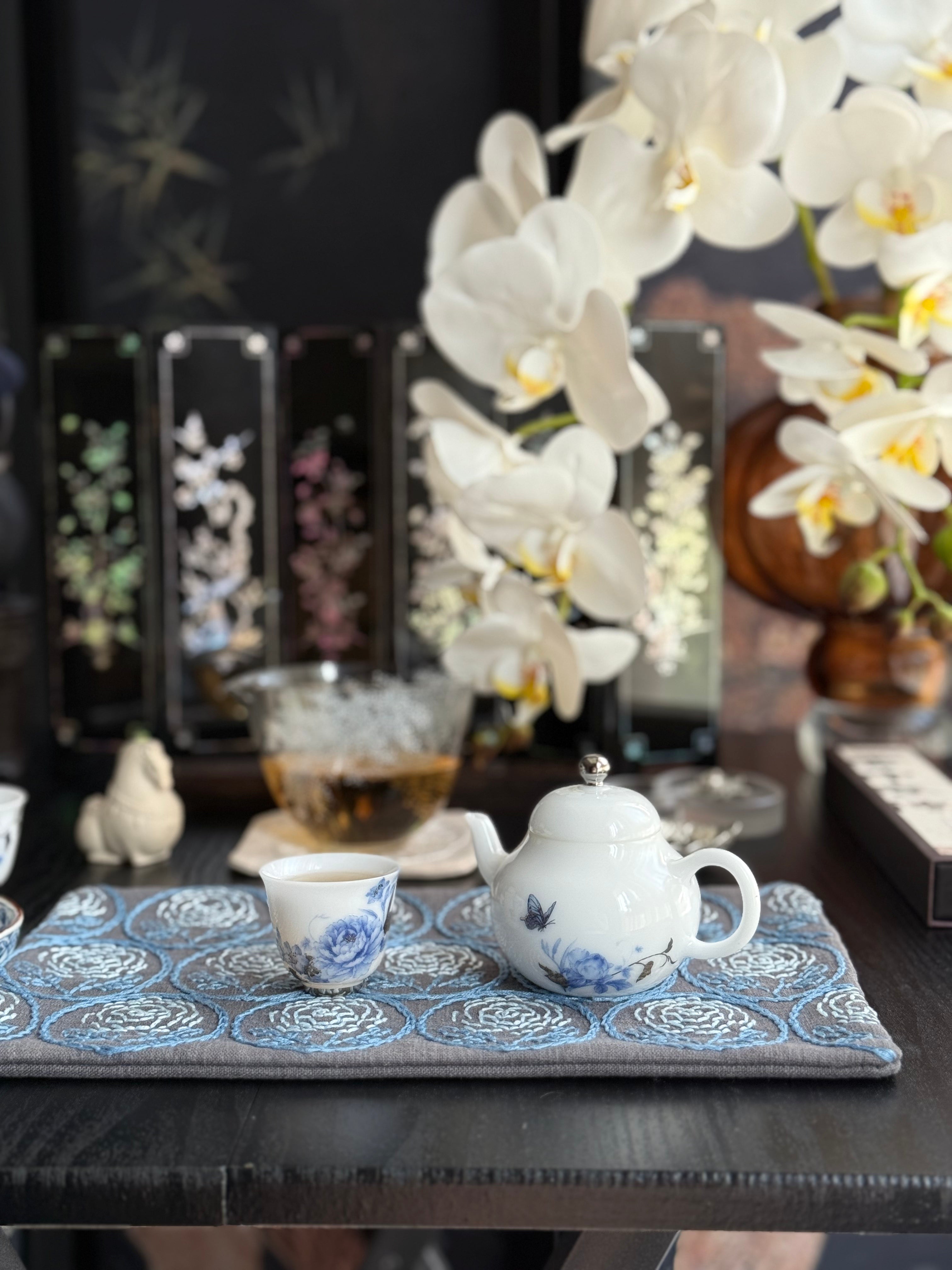 Tea set with a teapot and cups on a decorative mat, surrounded by white orchids.