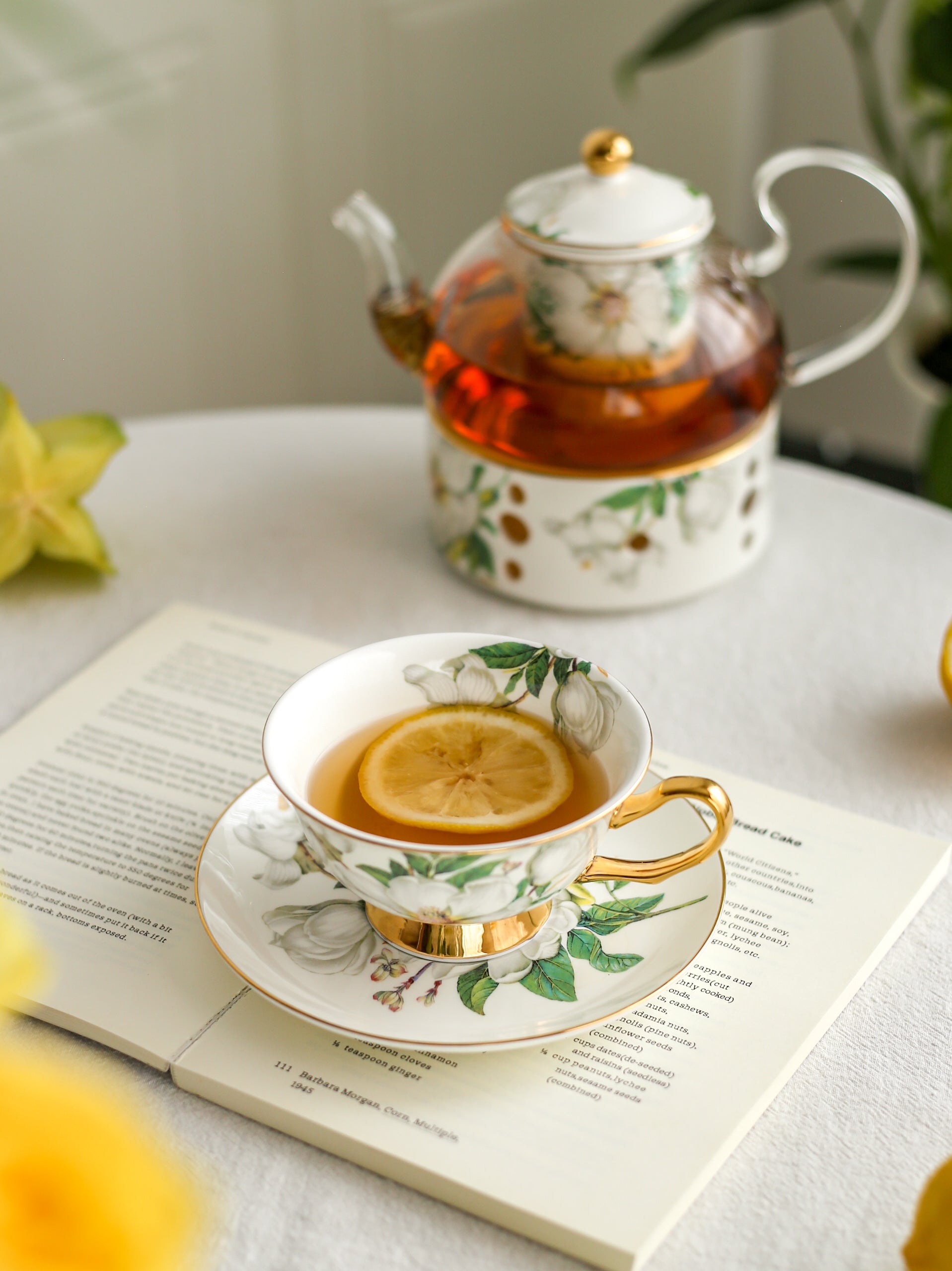 Tea cup with lemon slice on a saucer, teapot, and book on a table.