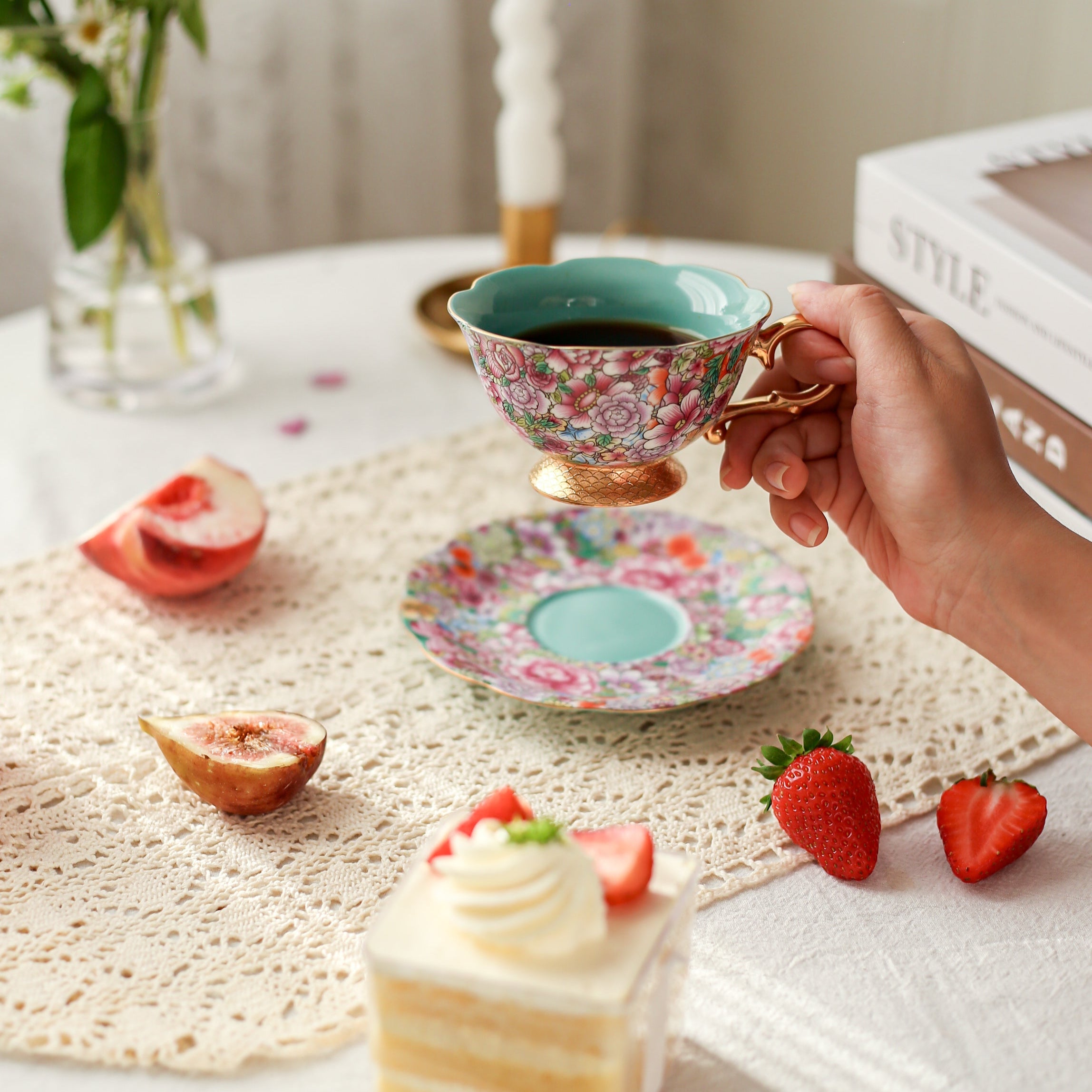 Hand holding a floral teacup with a slice of cake, strawberries, and flowers on a table.