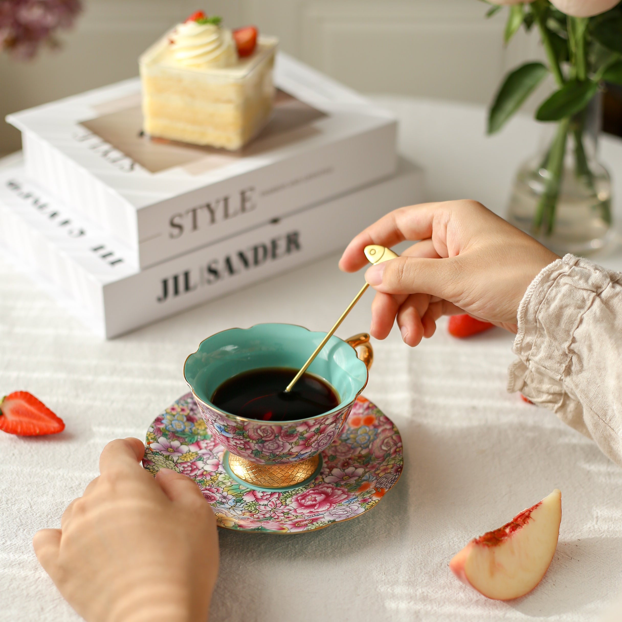 Person stirring coffee in a decorative cup with a cake and fruit on a table