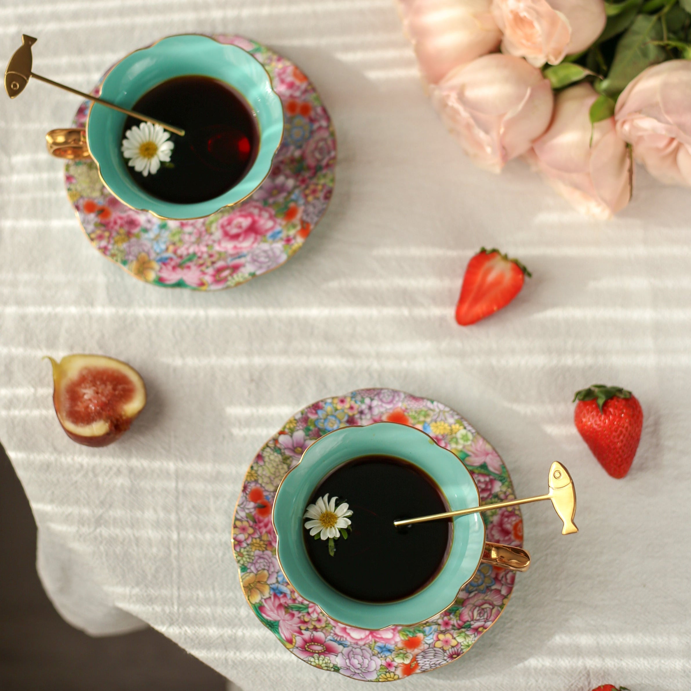 Two floral teacups with tea on a white tablecloth with fruit and flowers.