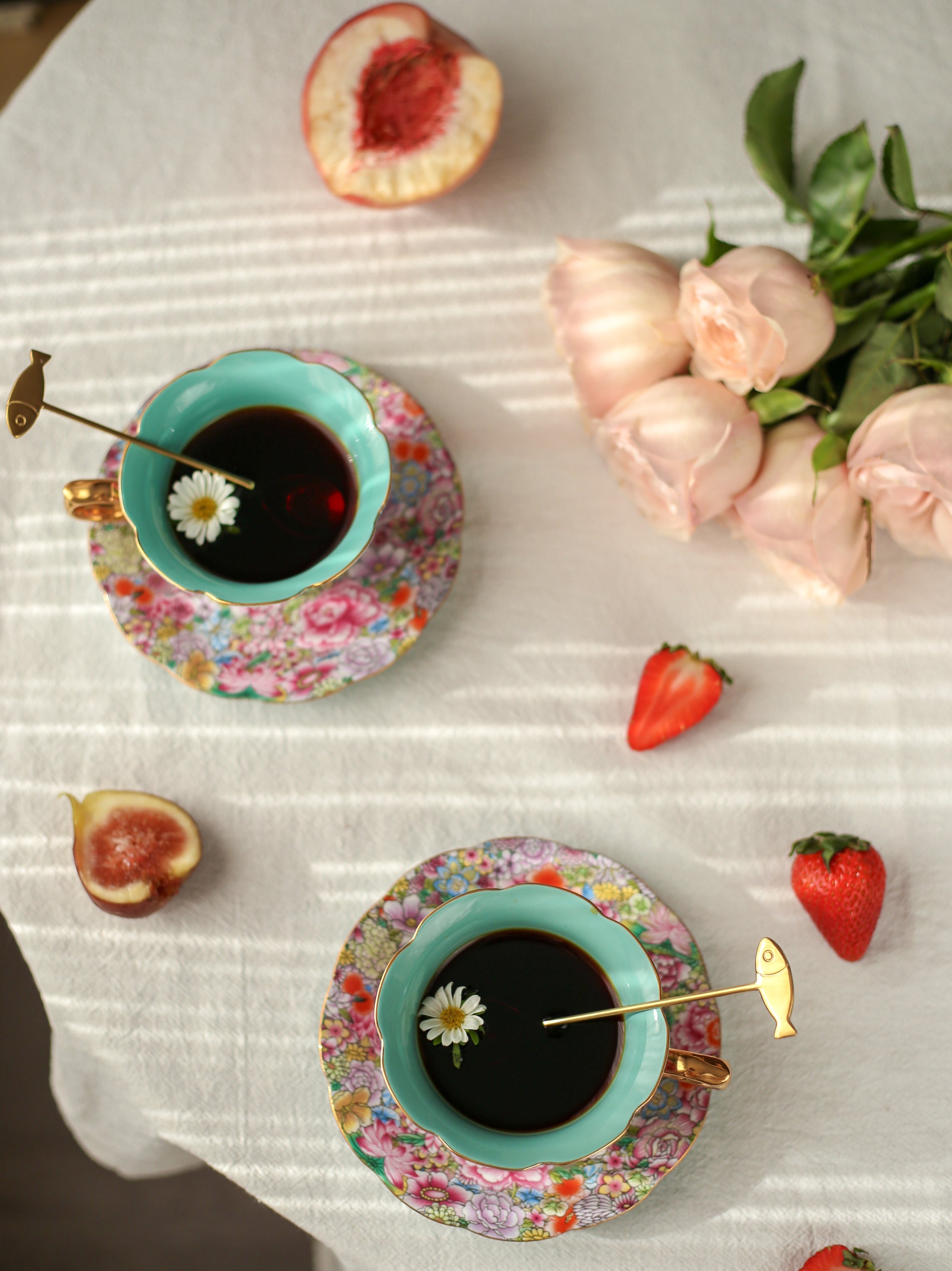 Two floral teacups with tea on a white tablecloth with fruit and flowers.