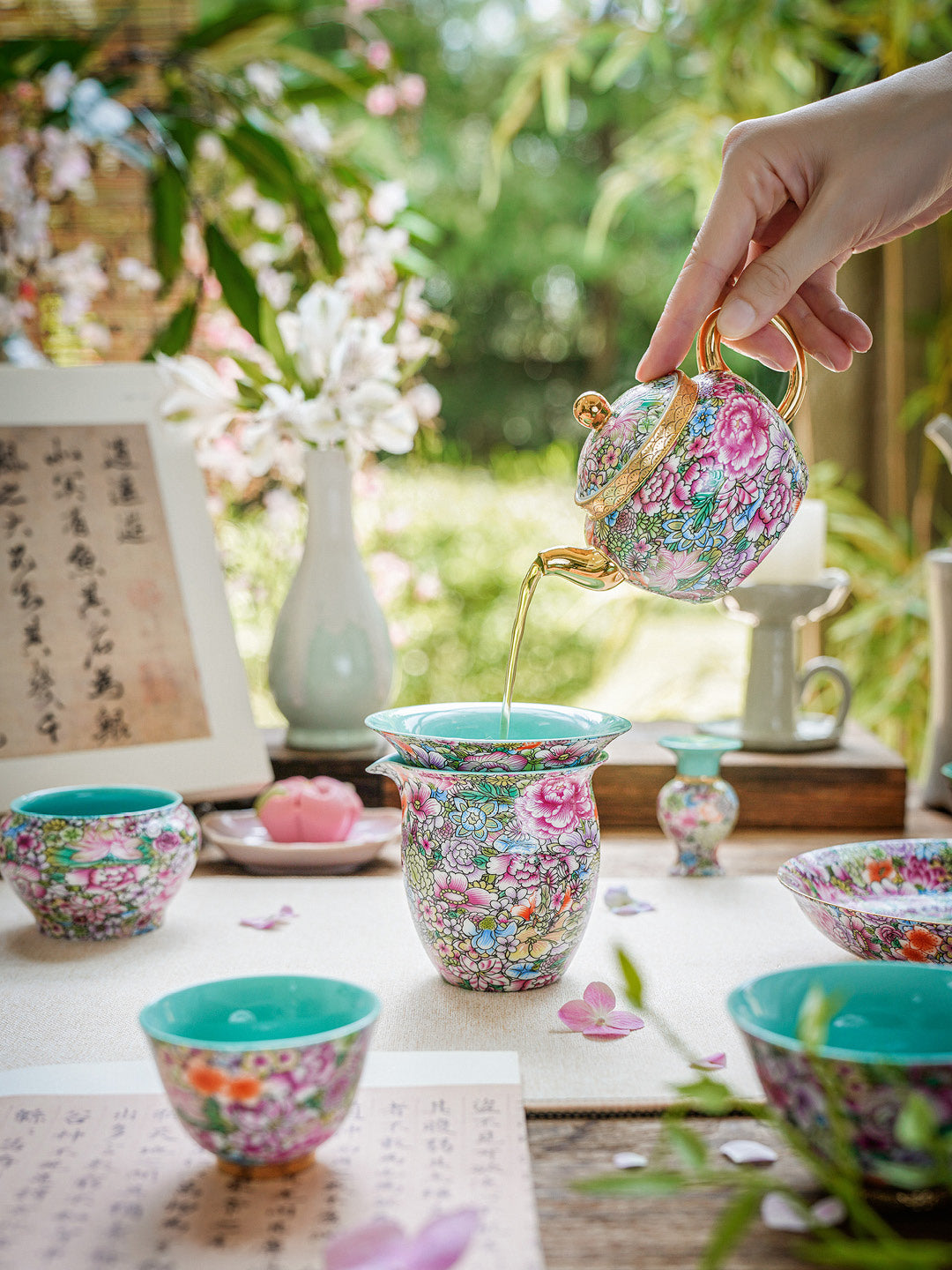 Person pouring tea from a floral teapot into a matching cup on a table with tea cups and a vase.