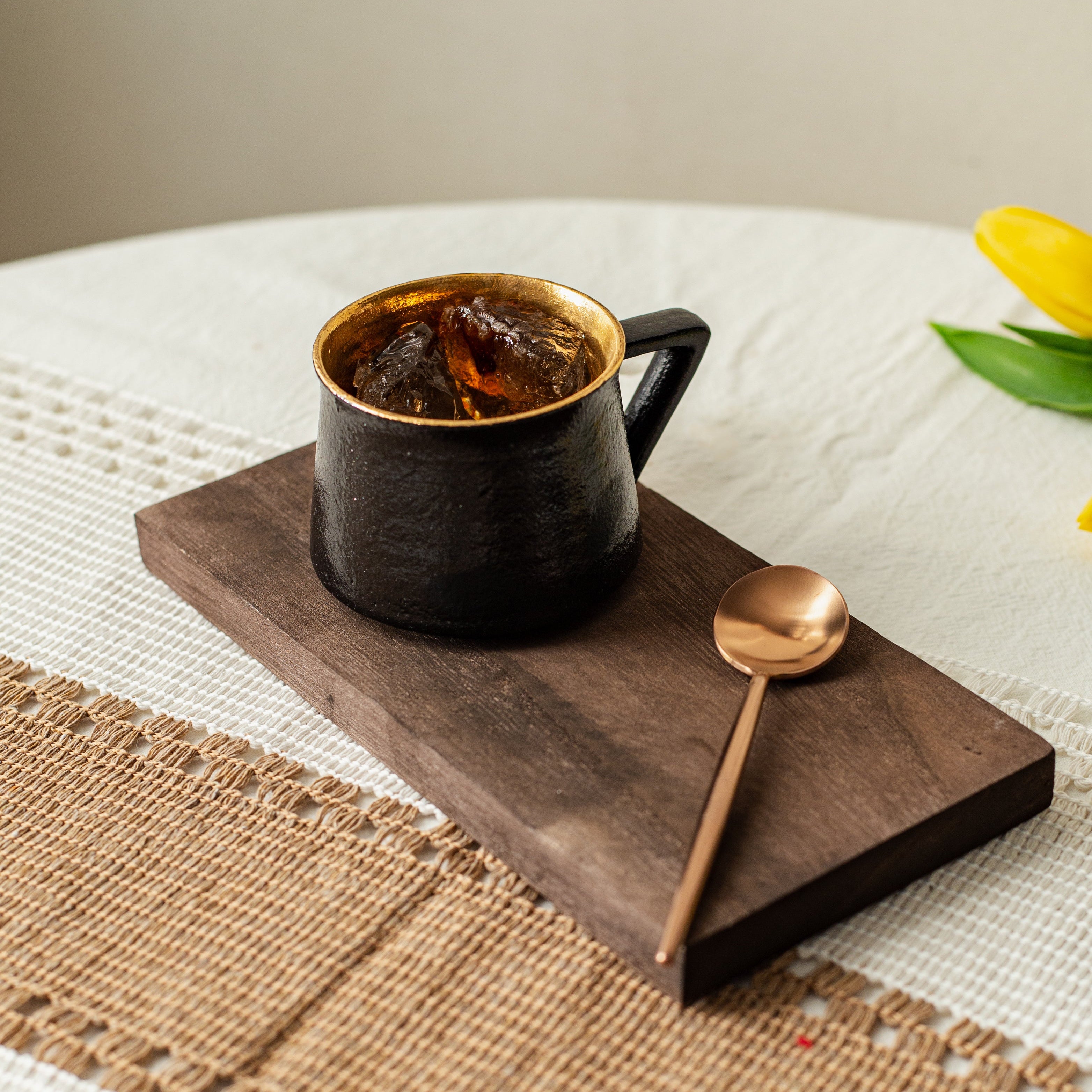 Black mug with gold interior on a wooden tray with a spoon, placed on a textured surface with a yellow flower.