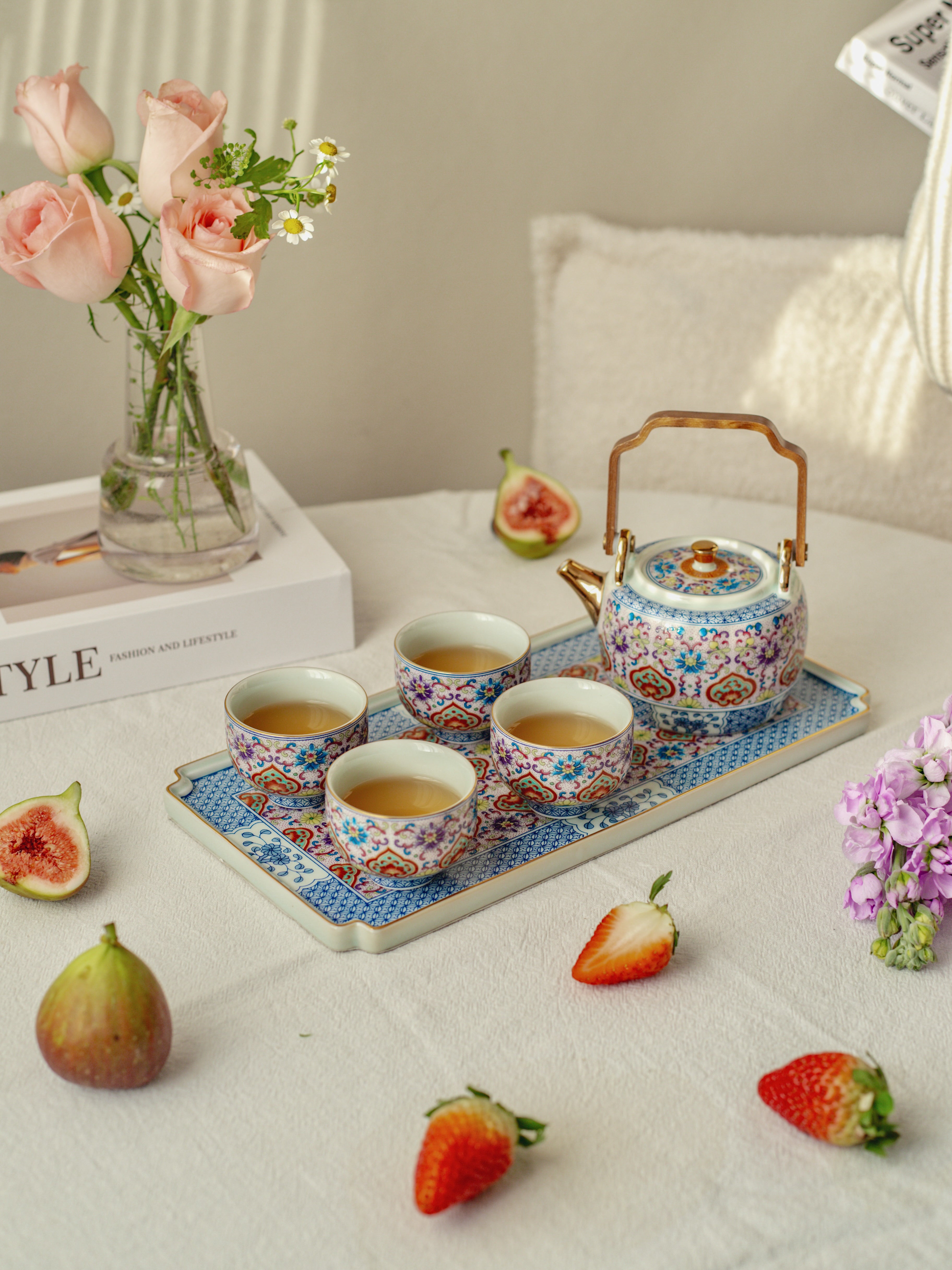 Tea set with teapot and cups on a tray, surrounded by strawberries and flowers on a table.