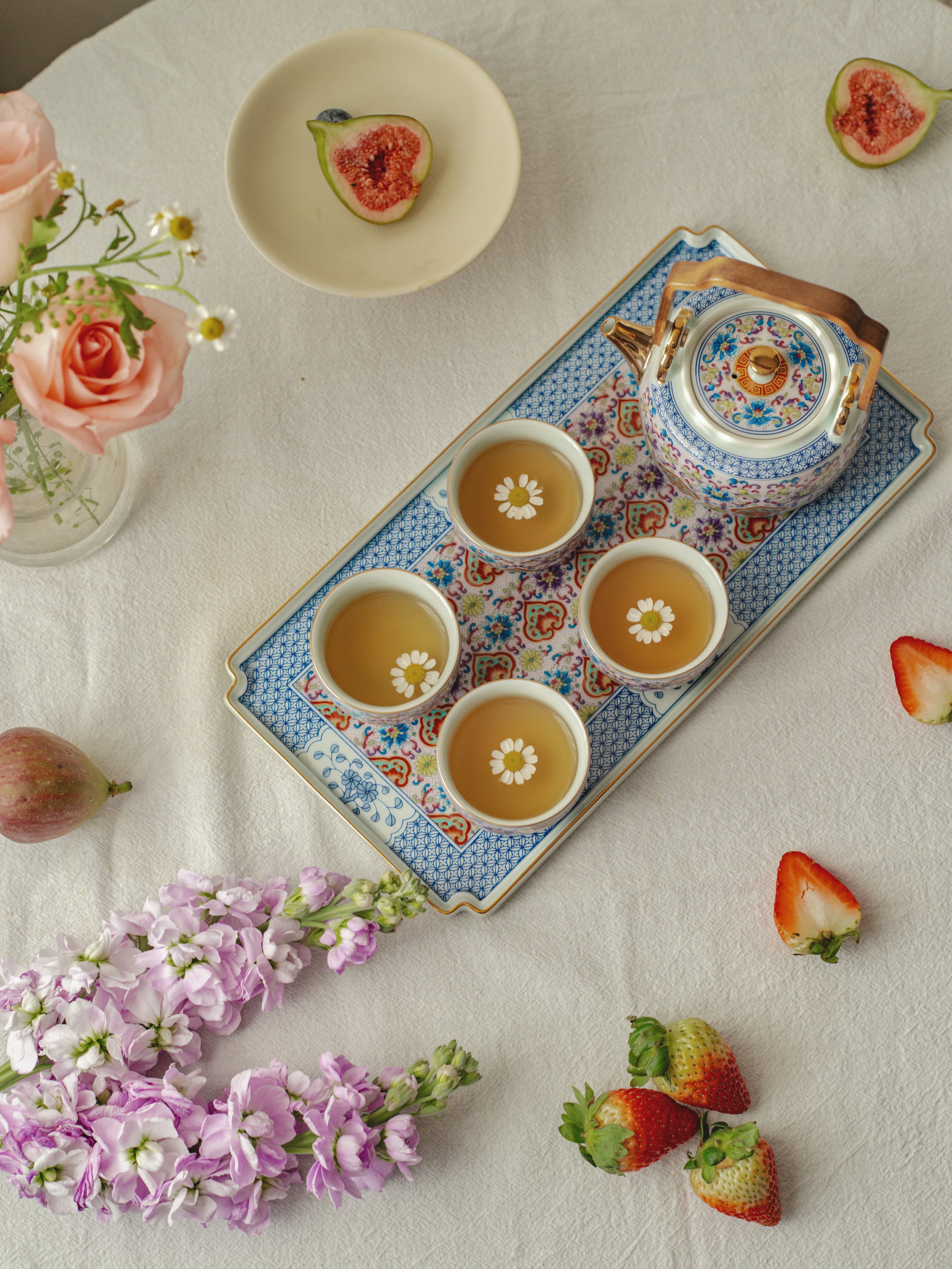 Tea set with cups and a teapot on a decorative tray, surrounded by flowers and fruits on a light surface.