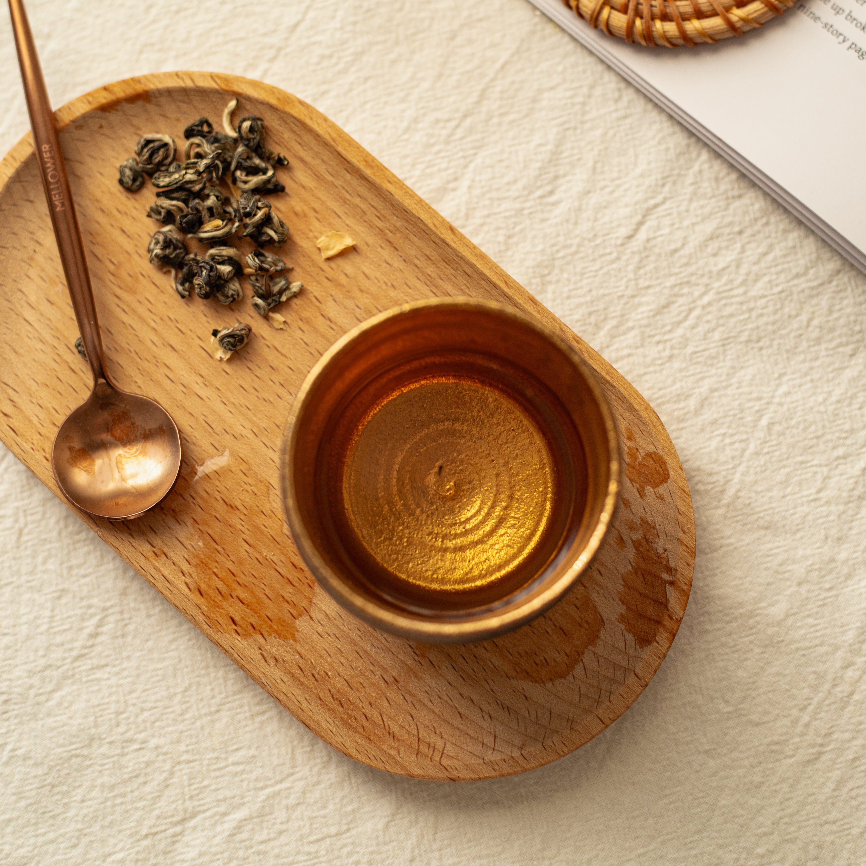 Brass cup on a wooden tray with tea leaves and a spoon, on a textured surface.