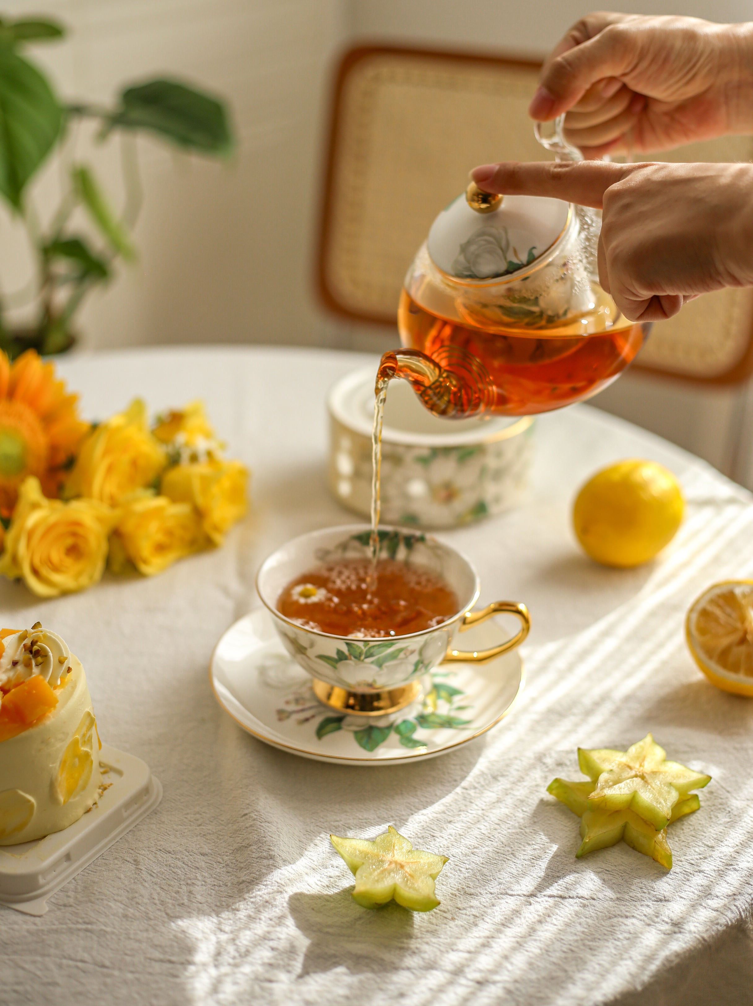 Person pouring tea from a teapot into a cup on a table with flowers and lemon slices.